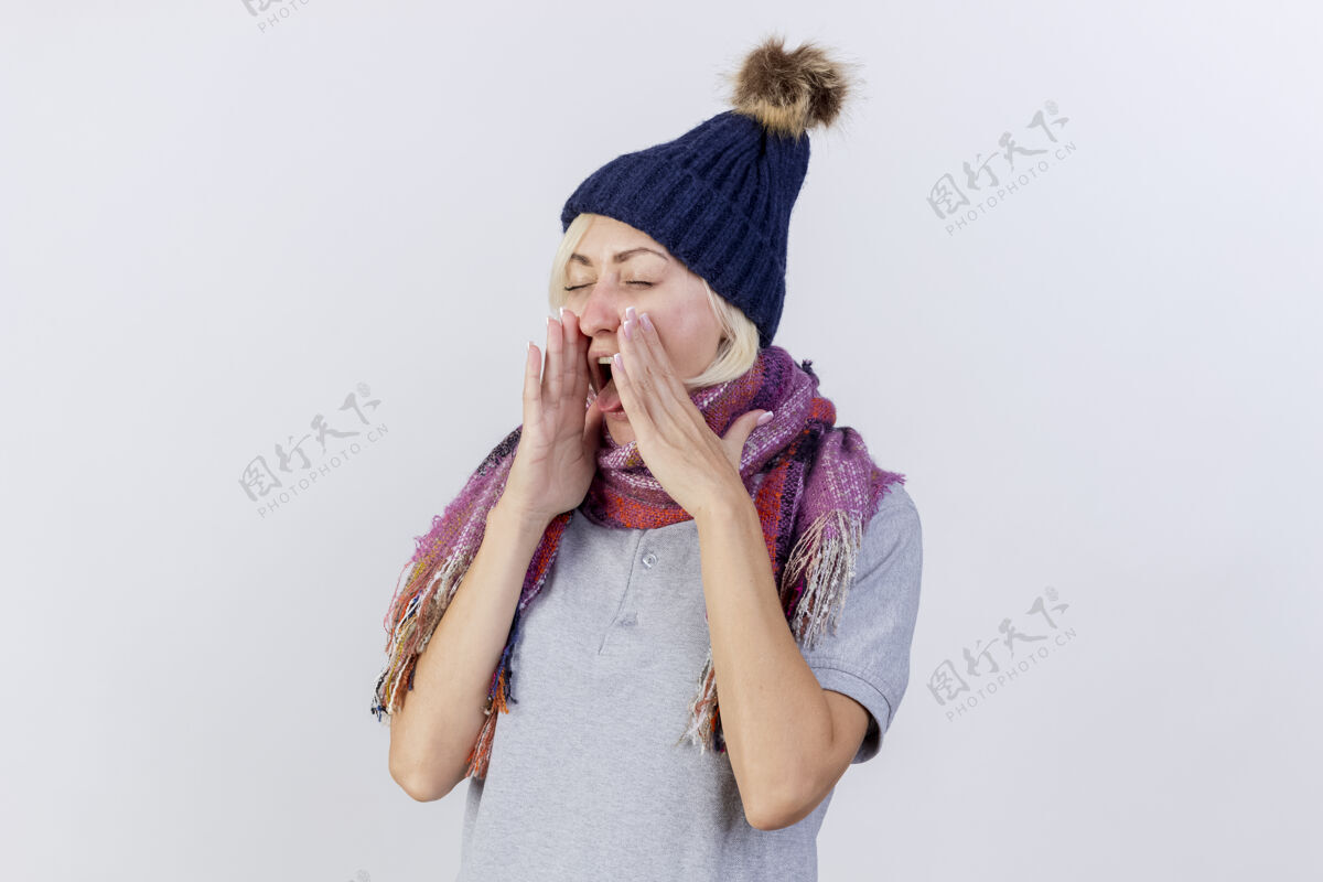 恼怒恼怒的年轻金发病女人戴着冬天的帽子和围巾结结巴巴地伸出舌头 双手紧握着隔离在白墙上的脸穿着冬天姿势