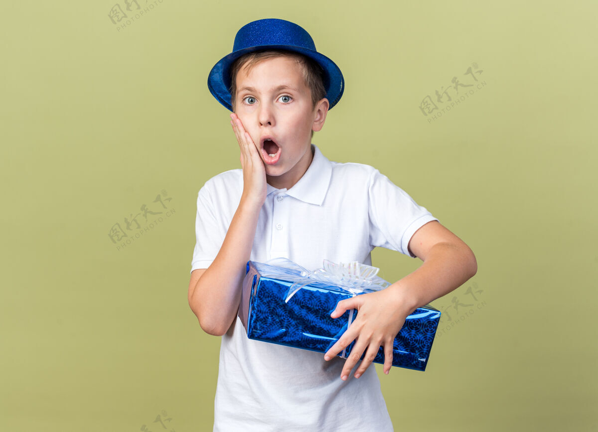 斯拉夫震惊的年轻斯拉夫男孩 戴着蓝色派对帽 手放在脸上 手拿着礼盒 孤立地放在橄榄绿的墙上 还有复制空间男孩橄榄脸