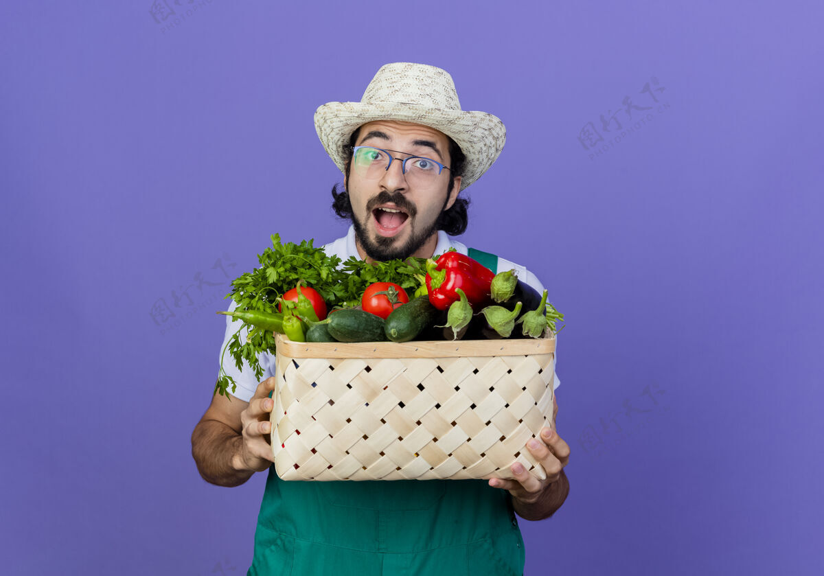 感觉年轻的留着胡须的园丁 穿着连身衣 戴着帽子 手里拿着装满蔬菜的板条箱 站在蓝色的墙上 高兴而兴奋地看着前方箱子胡须快乐