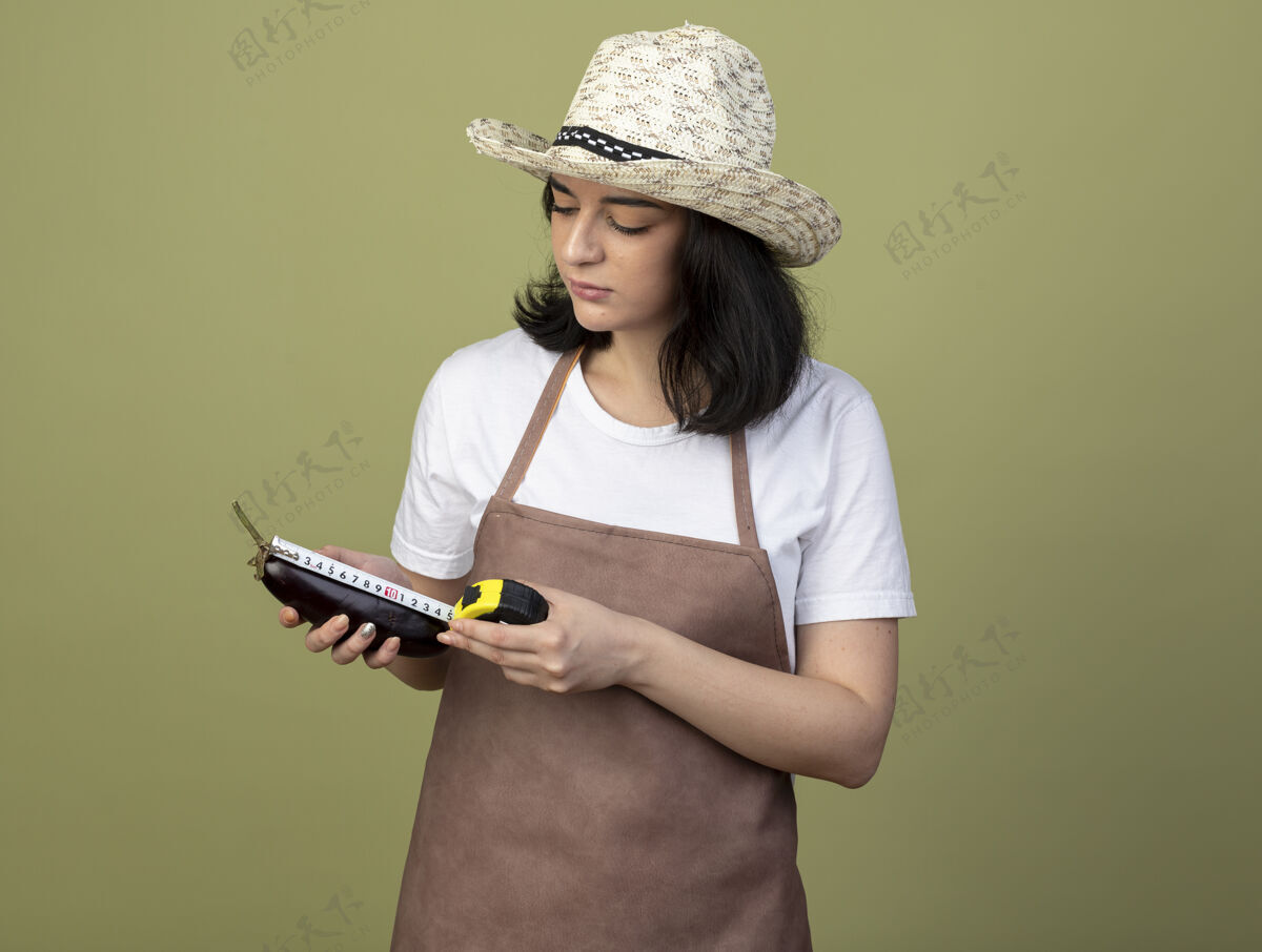 人自信的年轻黑发女园丁穿着制服戴着园艺帽用卷尺量茄子隔离在橄榄绿的墙上制服年轻人园艺