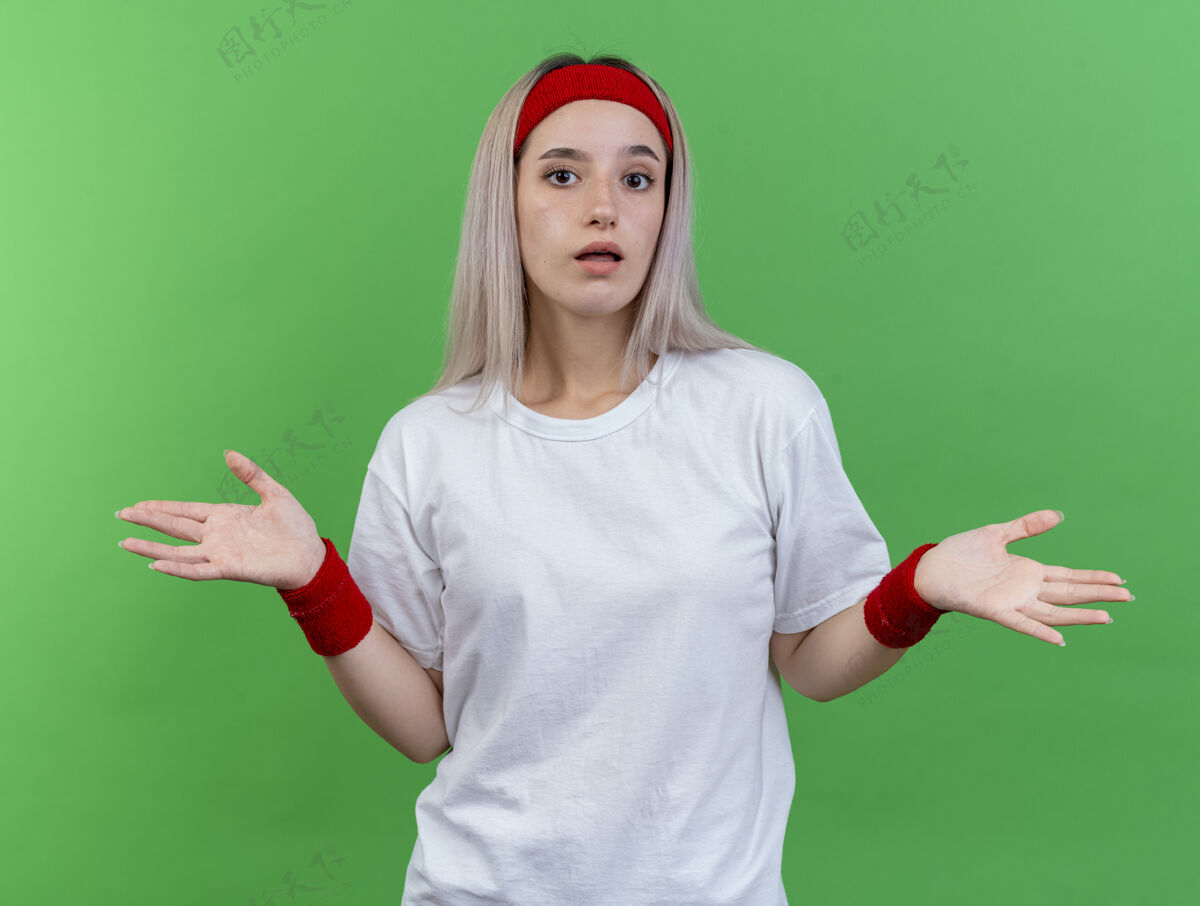 困惑困惑的年轻运动女性戴着背带 戴着头带和腕带 双手张开 孤立地站在绿色的墙上女性背带年轻