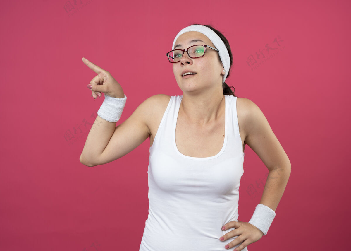 女人戴着眼镜 戴着头带和腕带的年轻运动女性 手放在腰上 手指孤立在粉红色的墙上人壁板人