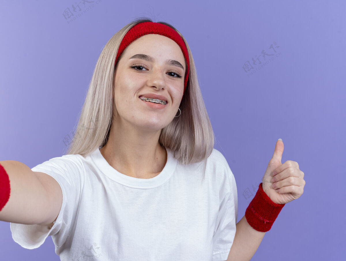 背带戴着头带和腕带的戴着背带的快乐的年轻运动女性竖起大拇指 看着紫色墙壁上孤立的前方请年轻人戴