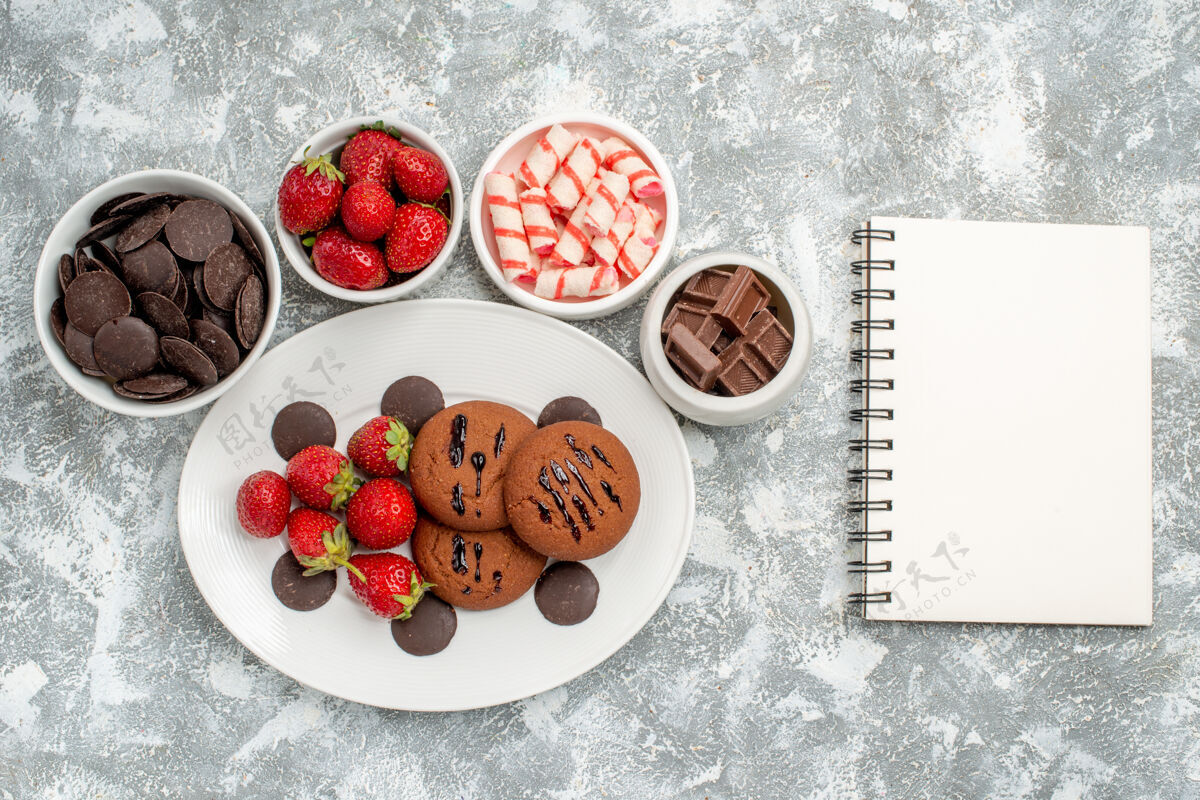 盘子俯瞰图：白色椭圆形盘子上的饼干 草莓和圆形巧克力 碗里放着糖果 草莓和巧克力 灰白色桌子上放着一本笔记本水果笔记本碗