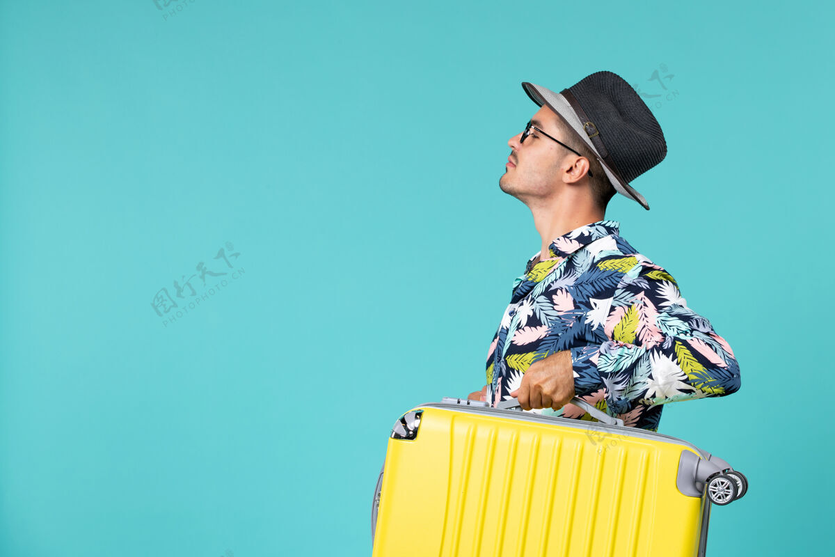 男人正前方的年轻男性准备旅行 并持有他的包在蓝色的墙上帽子举行人