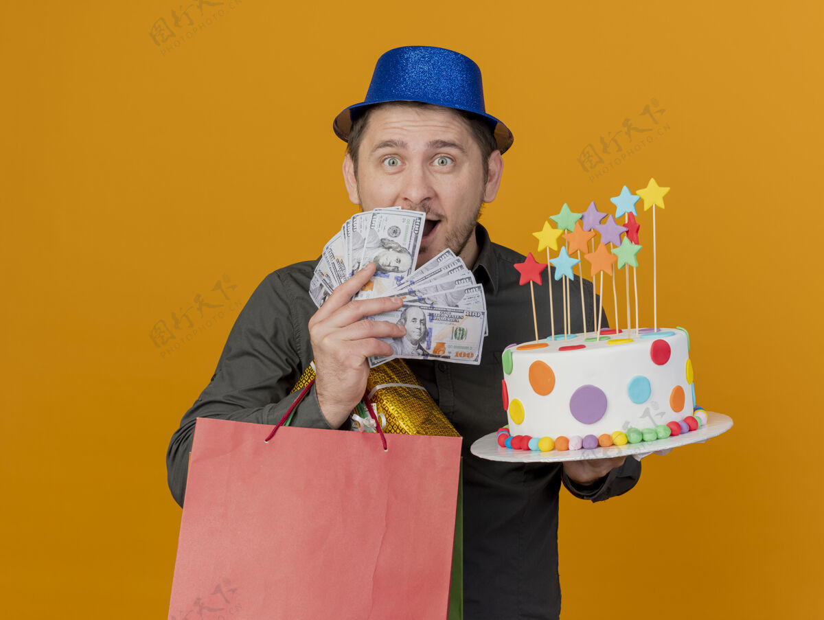 年轻人惊讶的年轻人戴着蓝色的帽子拿着礼物和蛋糕 脸上贴满了橘黄色的现金帽子穿上小伙子