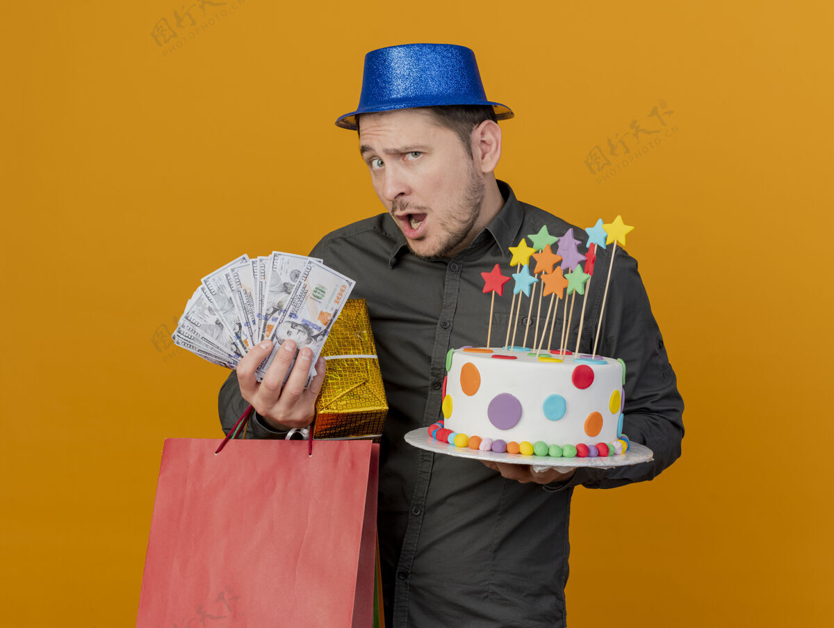 惊喜惊讶的年轻人戴着蓝色帽子拿着礼物蛋糕和现金孤立在橙色穿上年轻人橙色