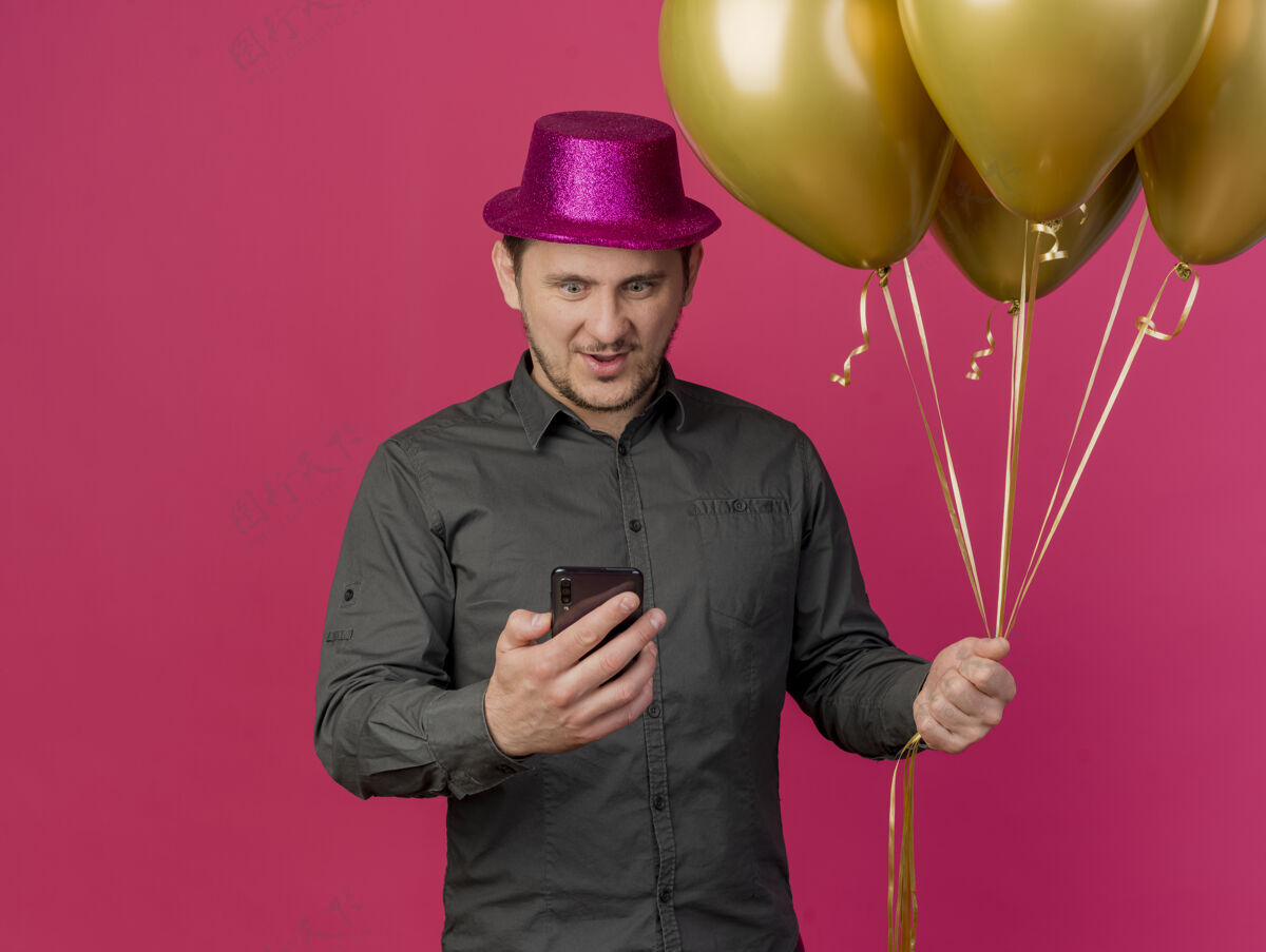 帽子惊讶的年轻人戴着粉红色的聚会帽 拿着气球 拿着手机 看着孤立的粉红色穿上惊喜派对
