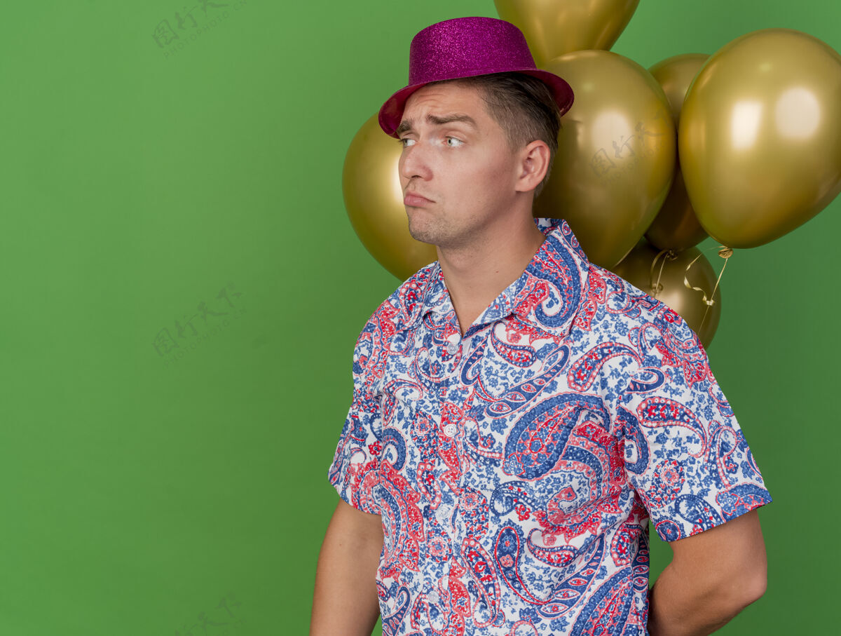 壁板悲伤的年轻人戴着粉红色的帽子站在绿色的气球前 看着旁边的聚会帽子站粉色