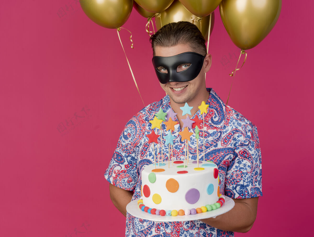 年轻人面带微笑的年轻人戴着化装眼罩站在气球前 手里拿着粉红色的蛋糕派对面具抱着