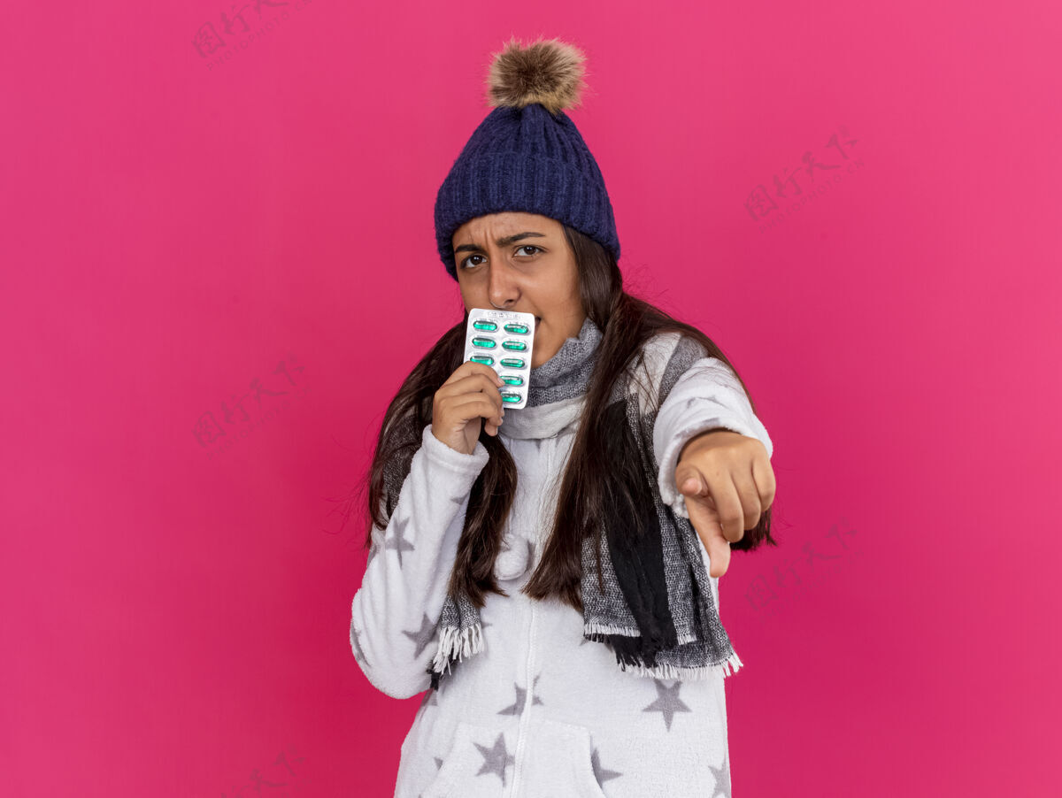 围巾严格的年轻生病的女孩戴着冬天的帽子和围巾 嘴里叼着药丸 显示你在粉红色上孤立的姿势戴着严格嘴巴