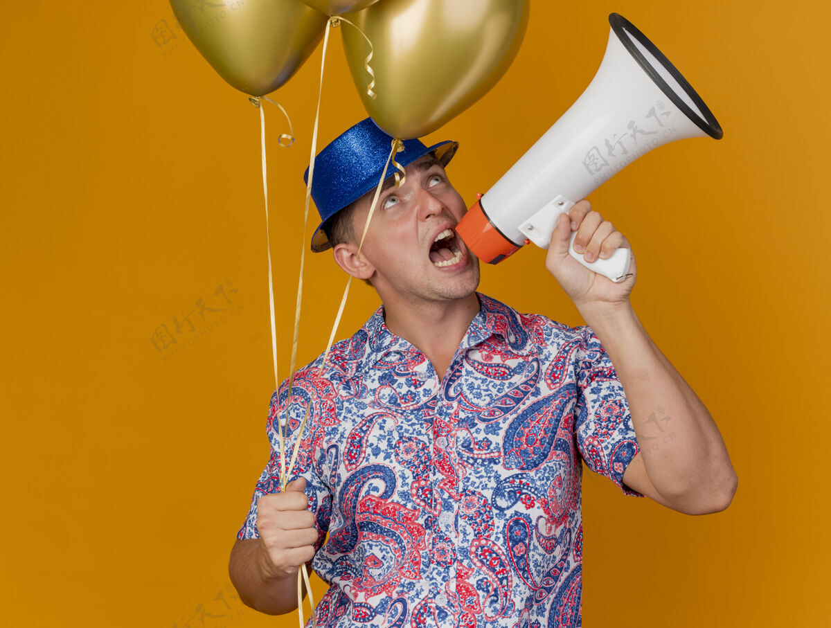 家伙快乐的年轻人戴着蓝色的帽子拿着气球在橙色的扬声器上讲话说话帽子派对