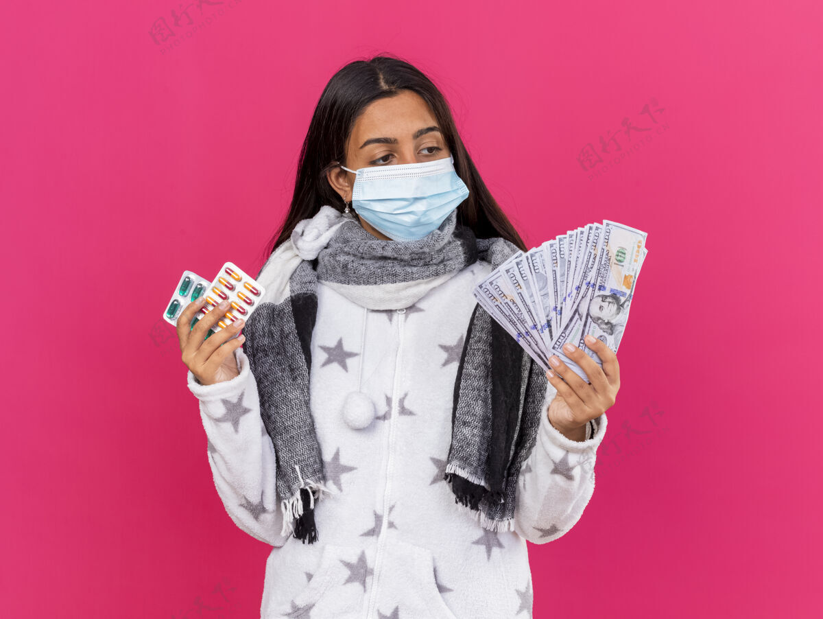 思考年轻生病的女孩戴着医用口罩戴着围巾拿着药片看着手里的现金孤立在粉红色的脸上药片佩戴疾病