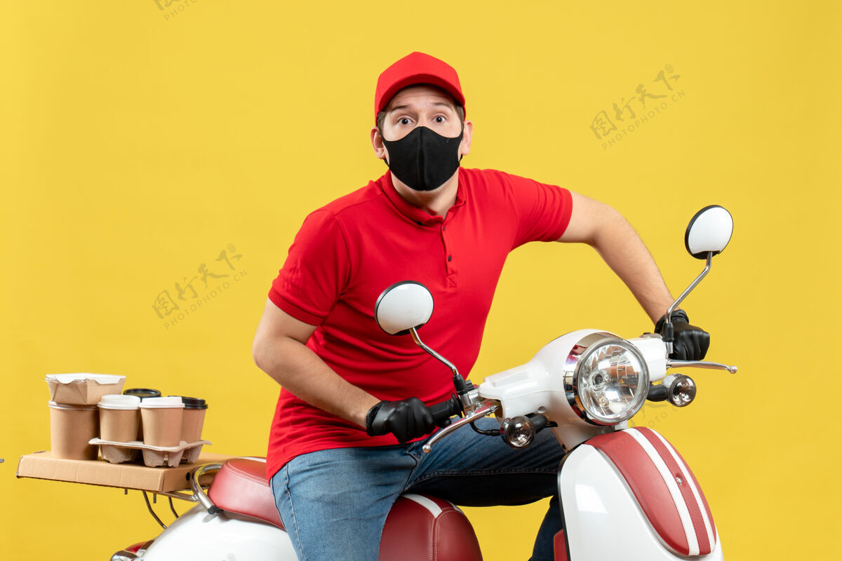 男性戴着医用面罩 穿着红色上衣和帽子手套的年轻人坐在黄色背景的踏板车上传递命令的俯视图男人背景送货