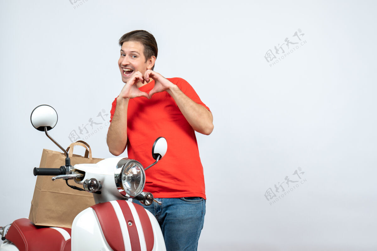 运动正面图是微笑的送货员 身穿红色制服 站在踏板车旁 在白色背景上做着心形手势送货员站立活跃