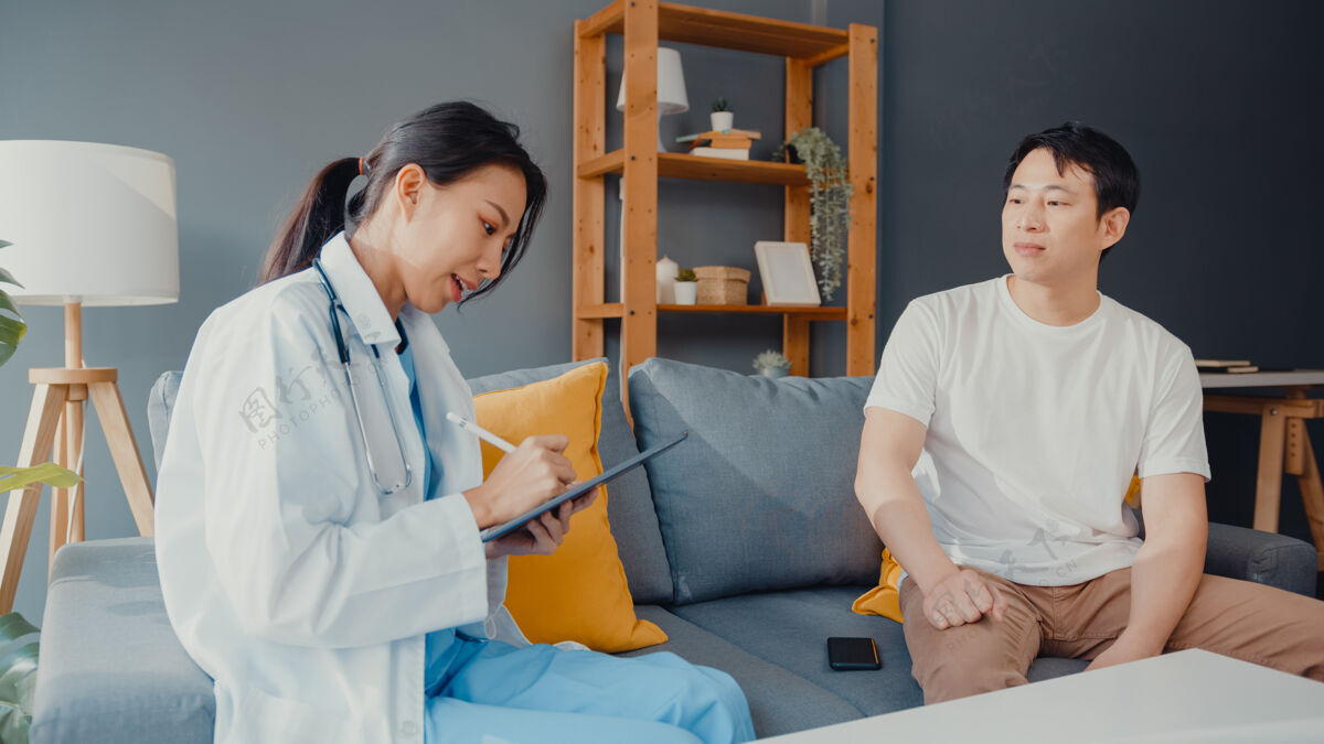 听诊器年轻的亚洲女专业医师医生用数字平板电脑和快乐的男病人分享健康测试的好消息坐在家里的沙发上访问诊所客厅