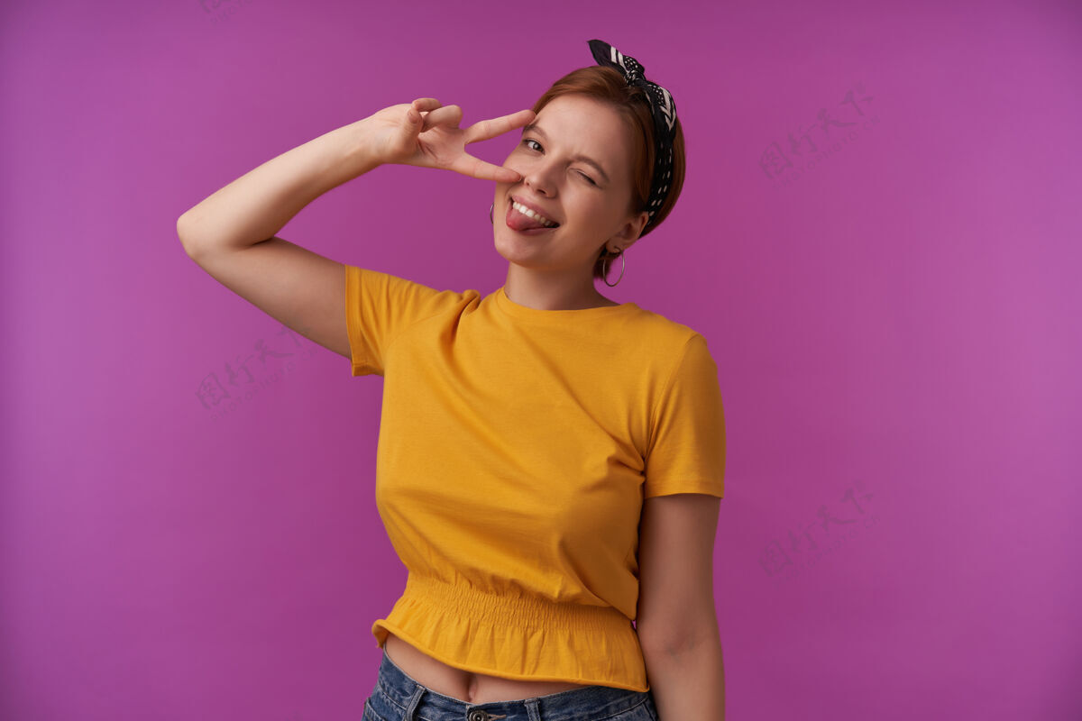 眨眼自然妆容的女人穿着黄色的时髦衬衫和黑色的大手帕 在紫色的墙上摆出两个手指在对你微笑情感调情眨眼高兴搞笑手女人年轻
