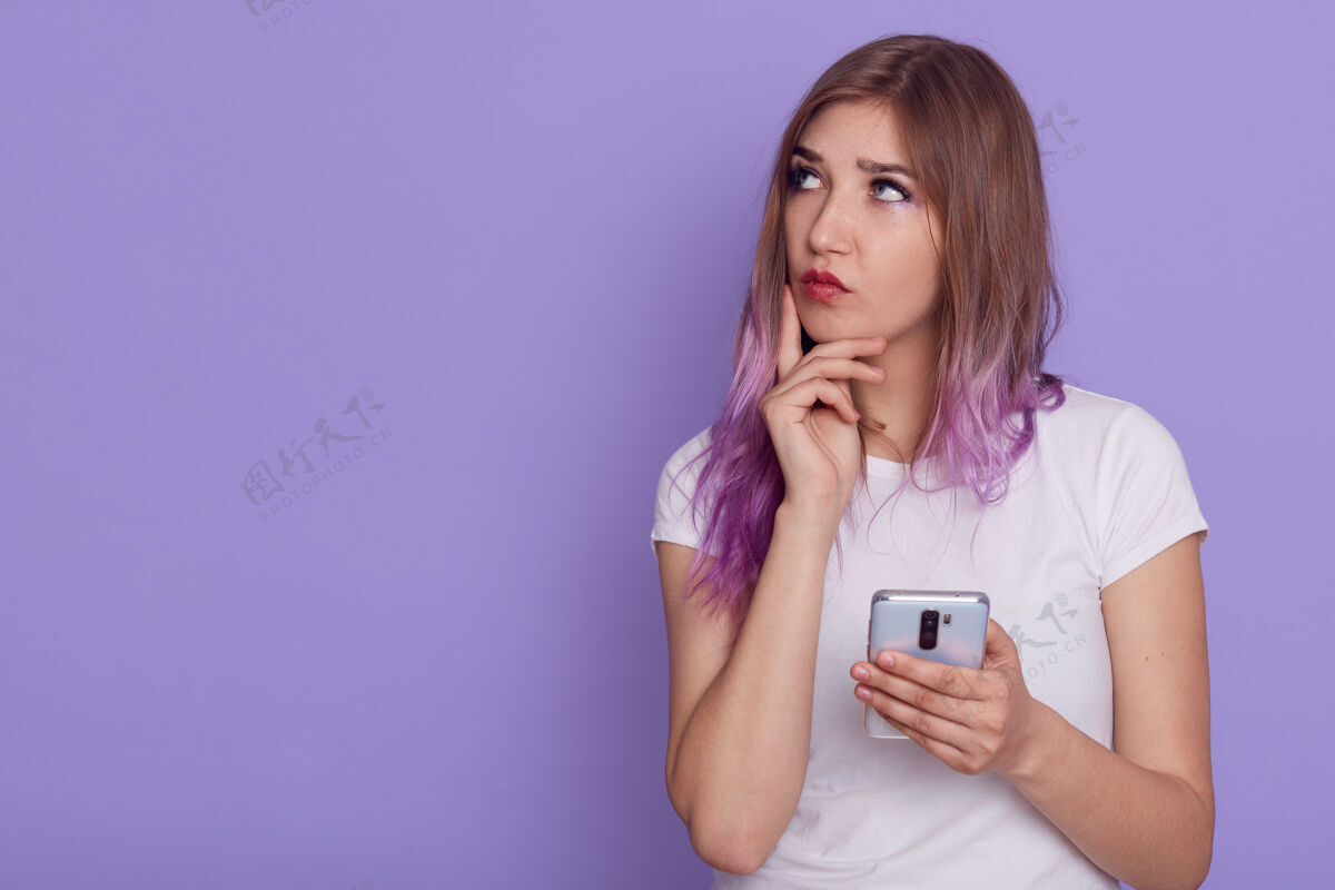 评论年轻可爱体贴的女人手里拿着智能手机 带着沉思的表情看着别处 手指贴在脸颊上 思考 复制空间 在紫色的墙上摆出孤立的姿势人信息聊天