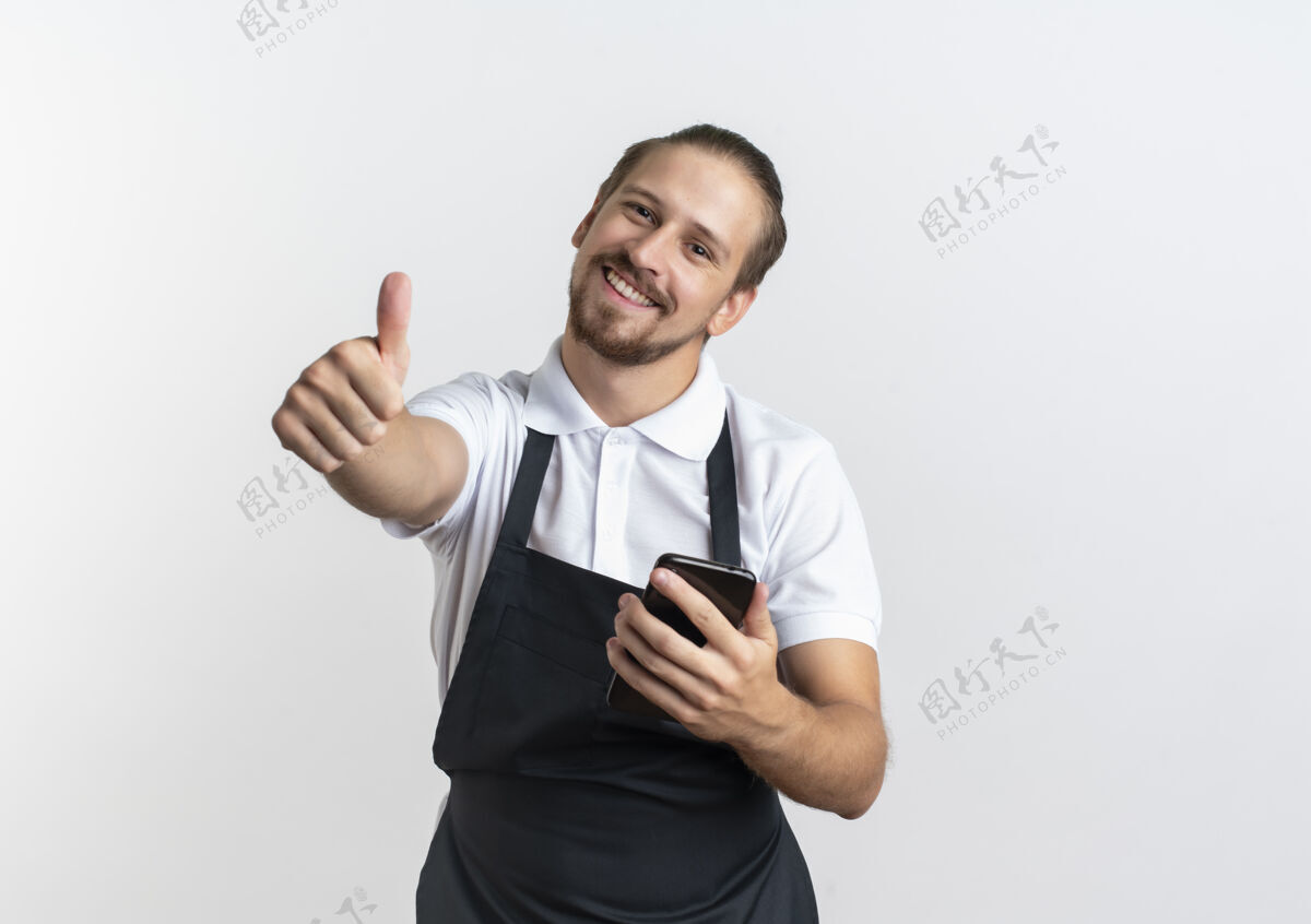 白面带微笑的年轻英俊的理发师穿着制服拿着手机 在白色的墙上孤立地竖起大拇指感觉抱着手机