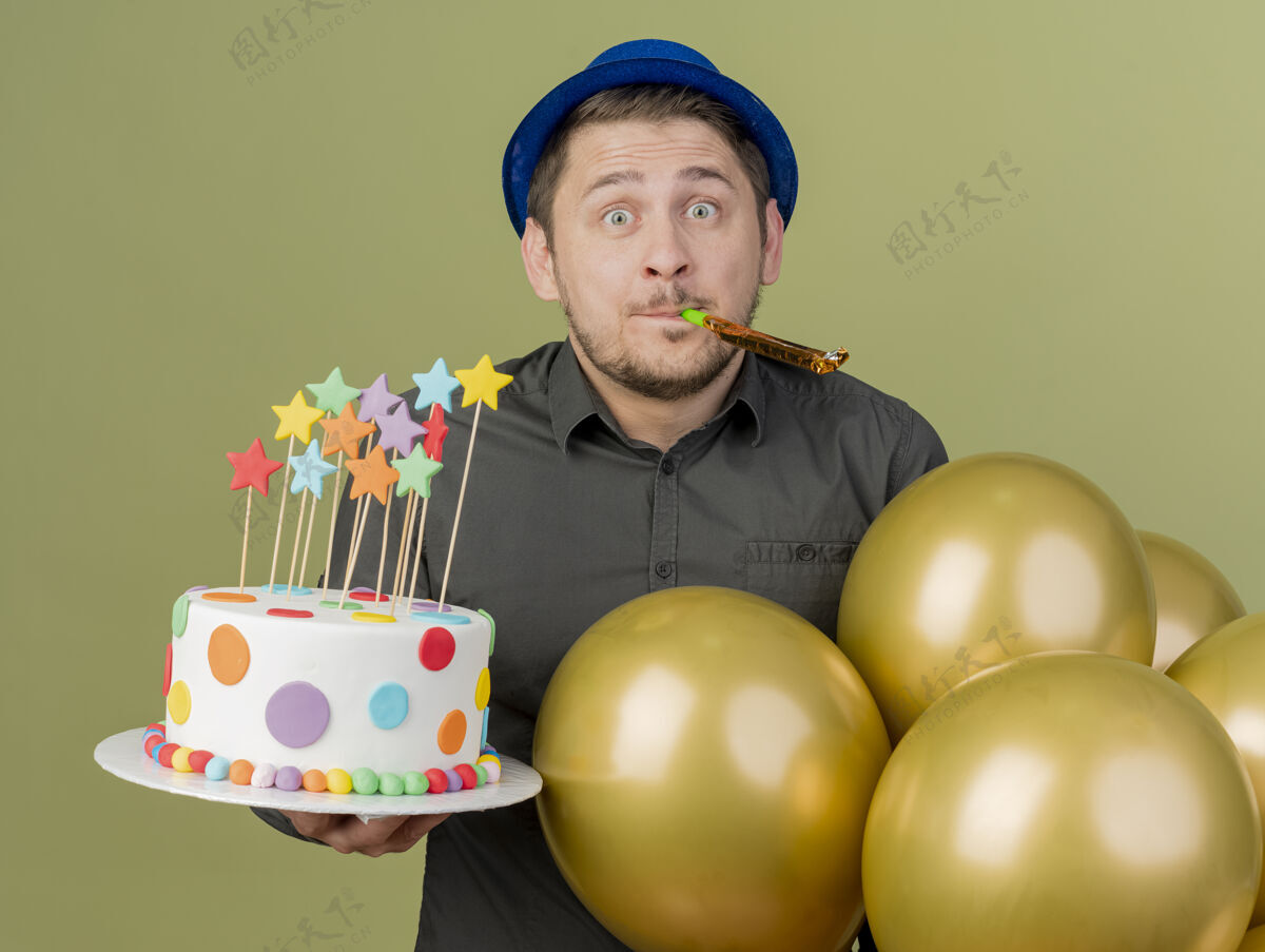 帽子惊讶的年轻人穿着黑色衬衫 戴着蓝色帽子拿着蛋糕和气球吹口哨孤立的橄榄绿橄榄绿色小伙子