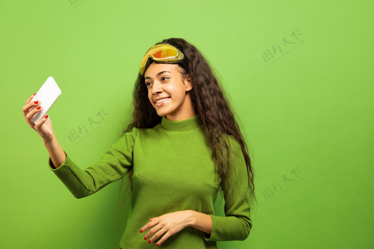 欢呼以绿色工作室为背景 黑色皮肤的非洲裔美国年轻女子滑雪面具肖像关于人类情感 面部表情 销售 广告 冬季运动和假期的概念自拍或虚拟博客微笑人手