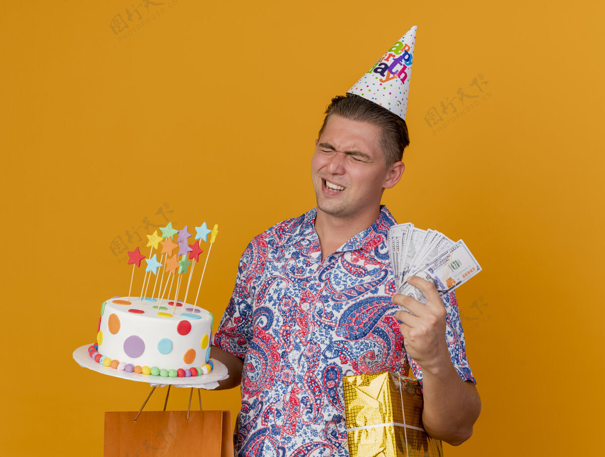 年轻年轻人戴着一顶闭着眼睛的生日帽 手里拿着礼物 蛋糕和现金被隔离在橘子上橙色穿封闭