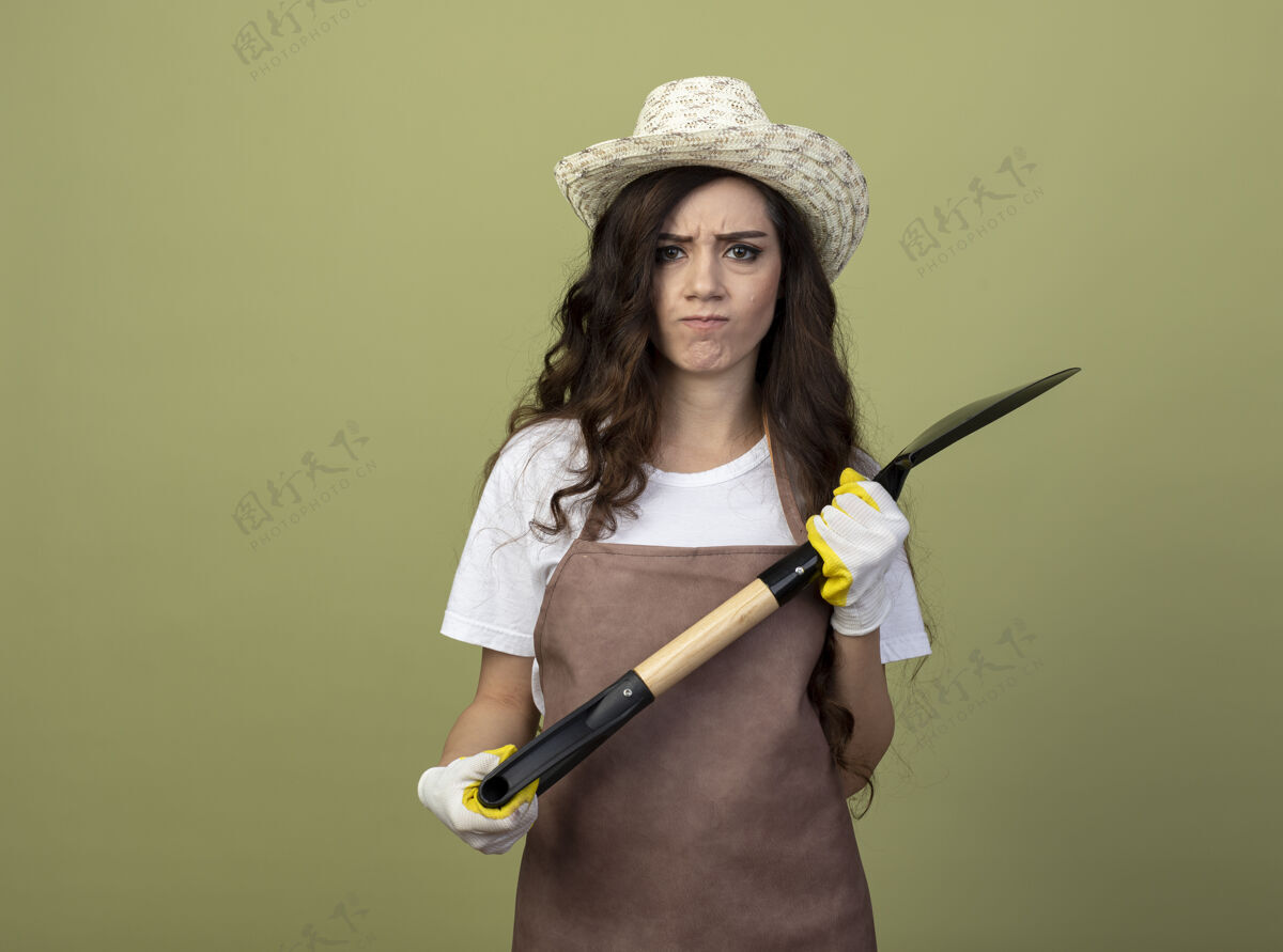 年轻穿着制服的年轻女园丁戴着园艺帽和手套 拿着一把铁锹孤立在橄榄绿的墙上园艺人表情