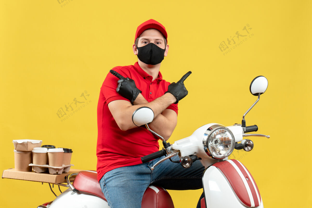 滑板车戴着医用口罩 穿着红色上衣 戴着帽子手套 坐在摩托车上 指着黄色背景的两边 思考的年轻人的正视图手套面罩命令