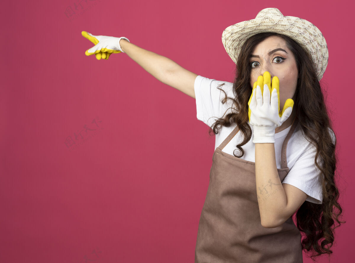 园艺震惊的年轻女园丁穿着制服 戴着园艺帽和手套 把手放在嘴上 并在粉红色的墙壁与复制空间隔离点的一边点壁板手套