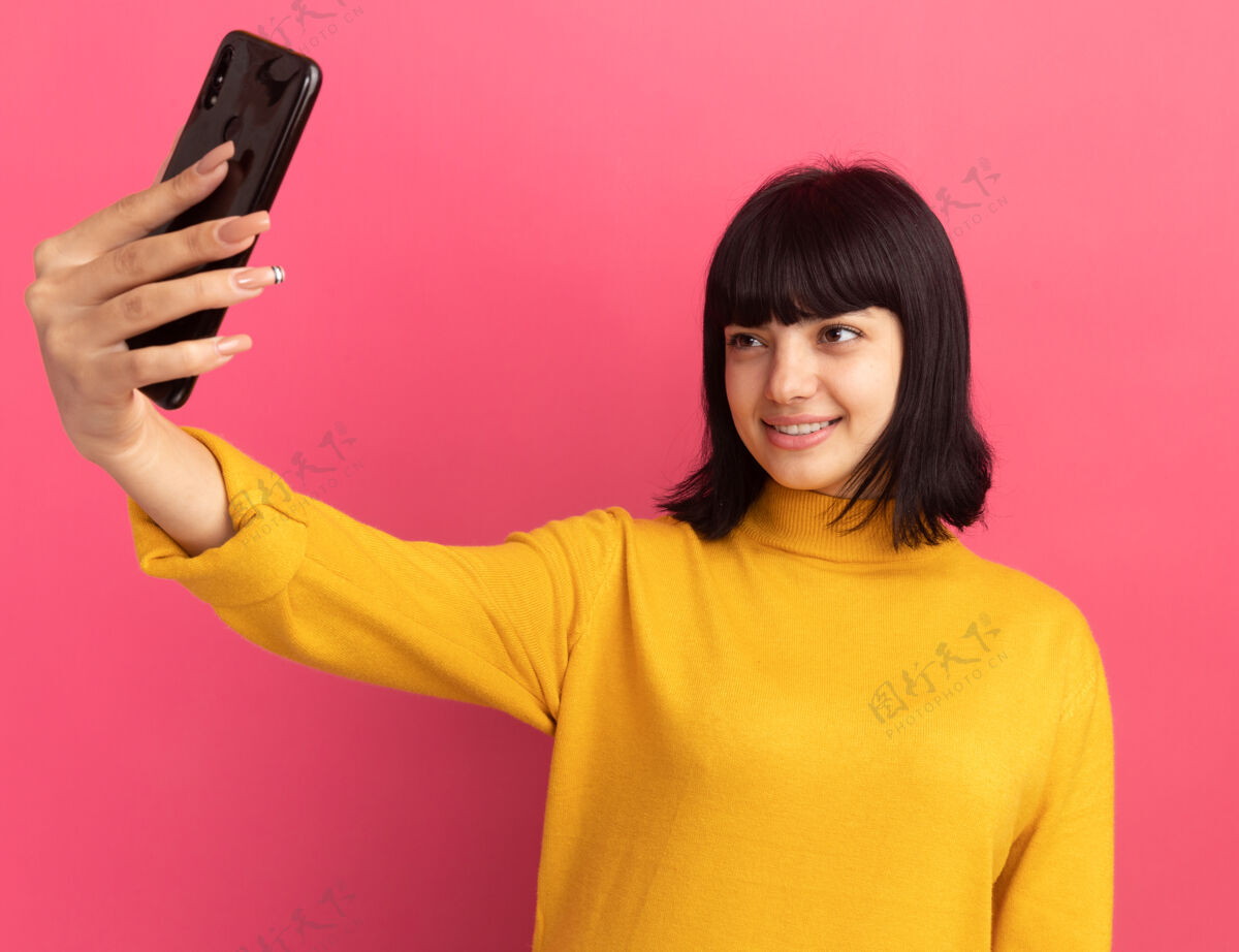 年轻的年轻的黑发白人女孩微笑着拿着手机 看着粉色的手机自拍黑发的拿着电话