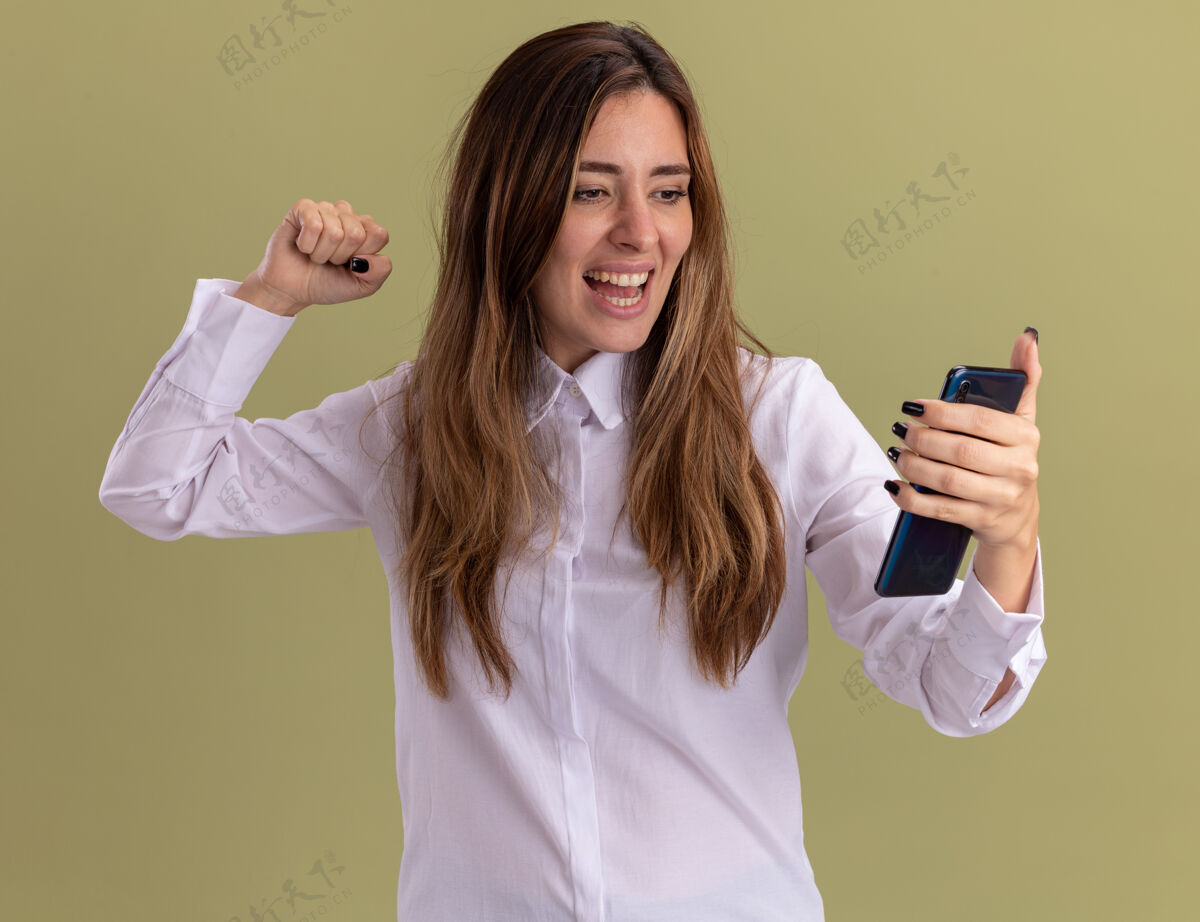 拳头快乐的年轻漂亮的白人女孩举起拳头站在那里 举着橄榄绿的手看着电话向上绿色年轻