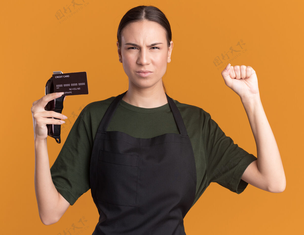保持恼怒的年轻黑发女理发师穿着制服 举起拳头拿着发夹和信用卡在橙色举行理发师头发