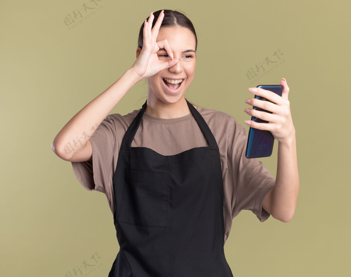 手指身着制服的快乐的年轻黑发理发女孩手持橄榄绿的手指看着电话理发师绿色电话