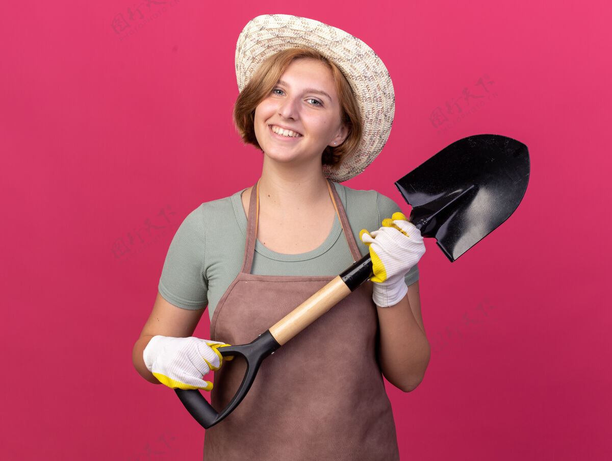 年轻微笑着的年轻斯拉夫女园丁戴着园艺帽和手套 手里拿着粉红色的铲子花园黑桃帽子