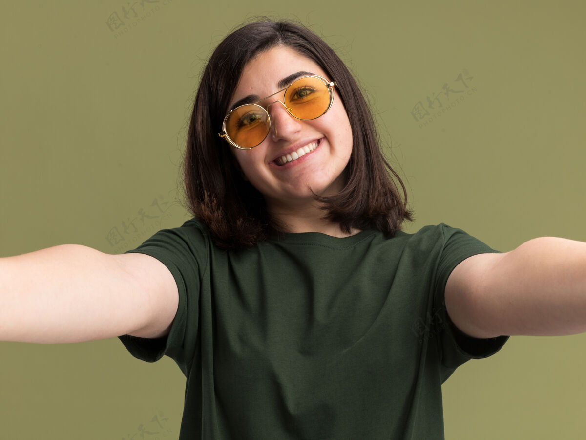 漂亮戴着太阳眼镜的年轻漂亮白人女孩微笑着假装拿着相机在橄榄绿上自拍拿着相机眼镜
