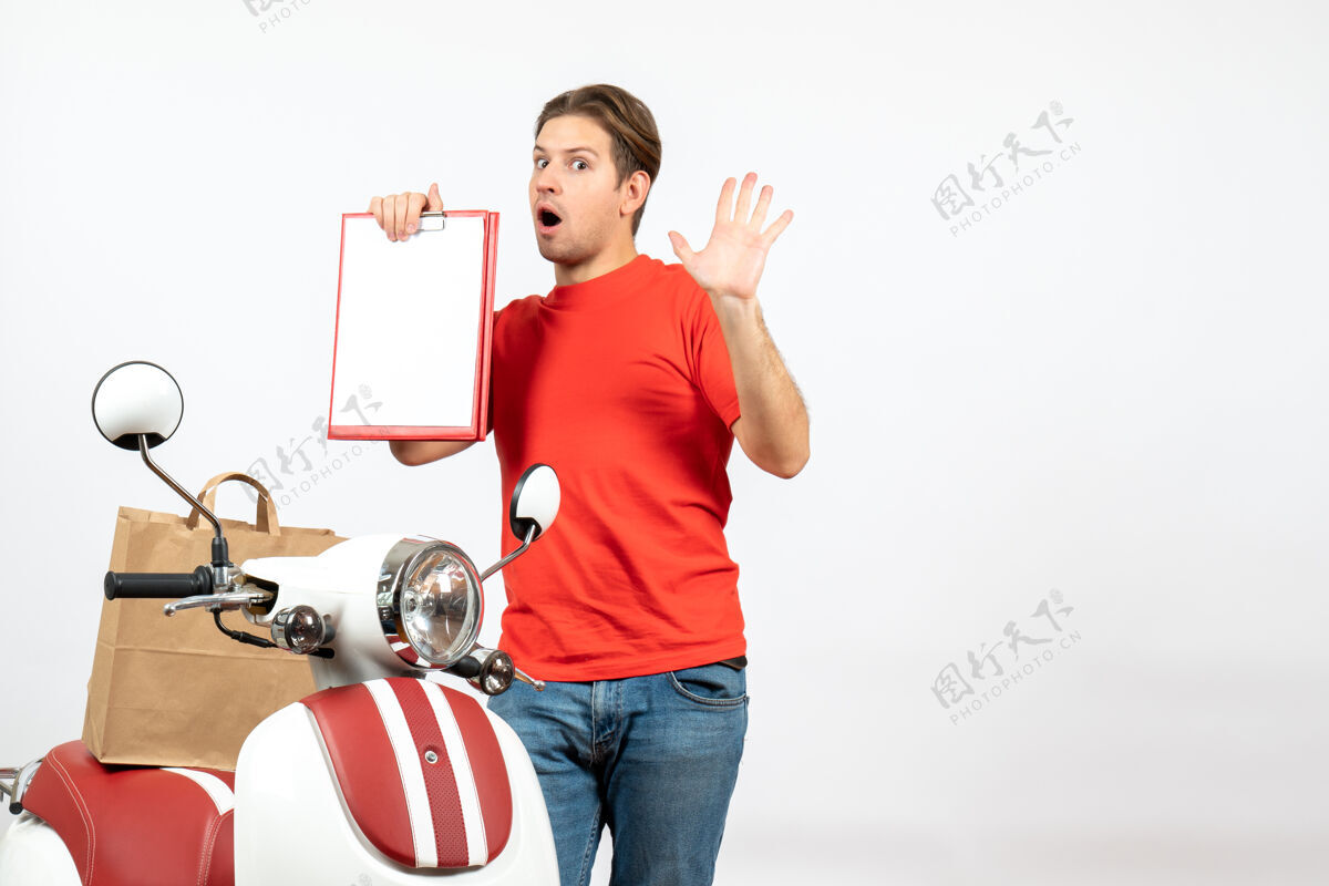 男性俯视图：身穿红色制服的年轻送货员站在滑板车旁 在白墙上展示文件表演送货员情绪