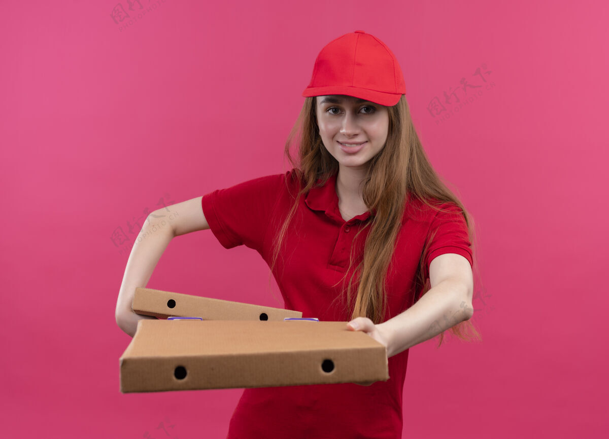 送货微笑着的年轻送货女孩穿着红色制服 拿着包裹 在孤立的粉色空间里伸展伸展年轻抱着