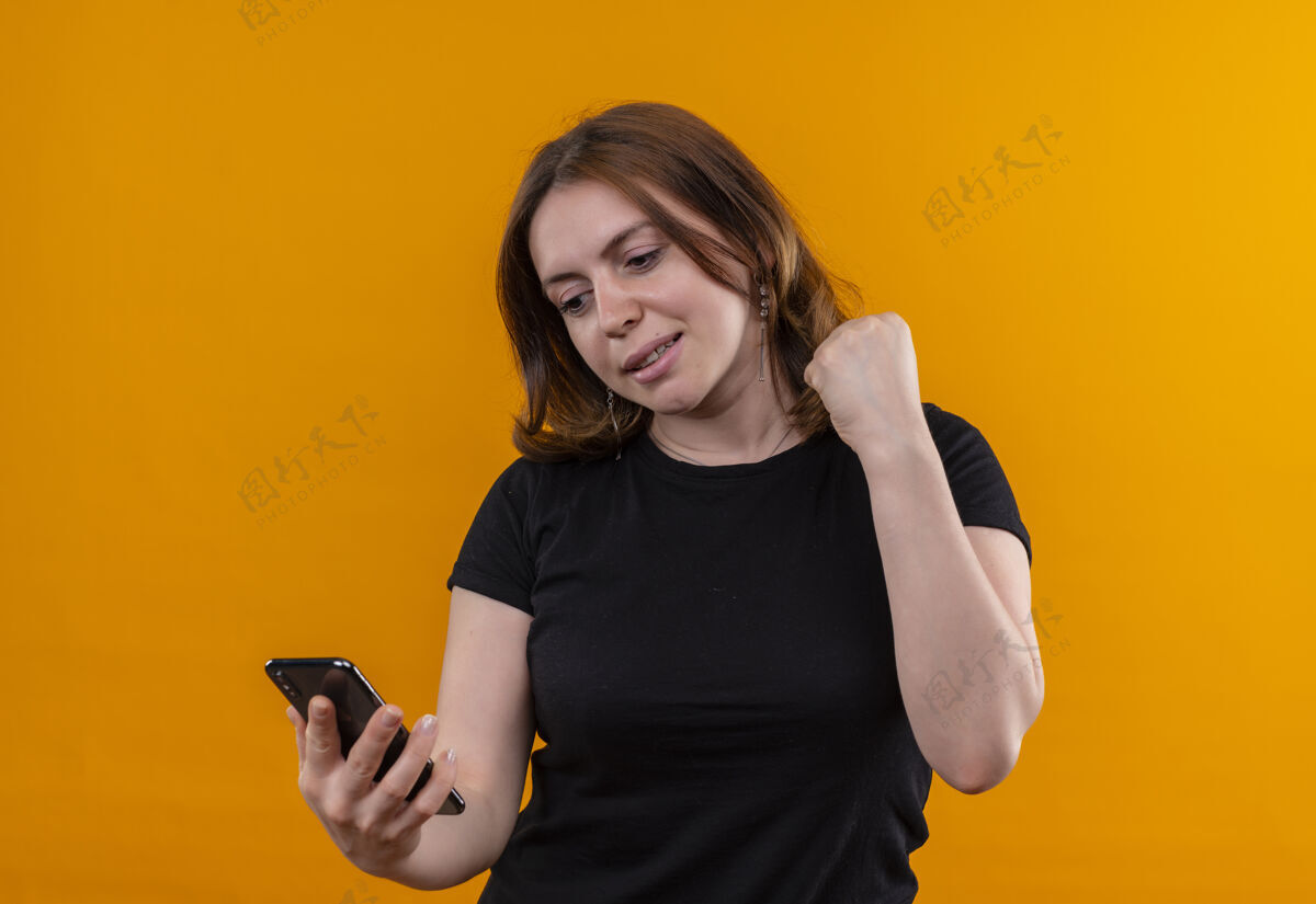微笑微笑的年轻休闲女子手持手机 在孤立的橙色空间举起拳头看着它橙色手机女人