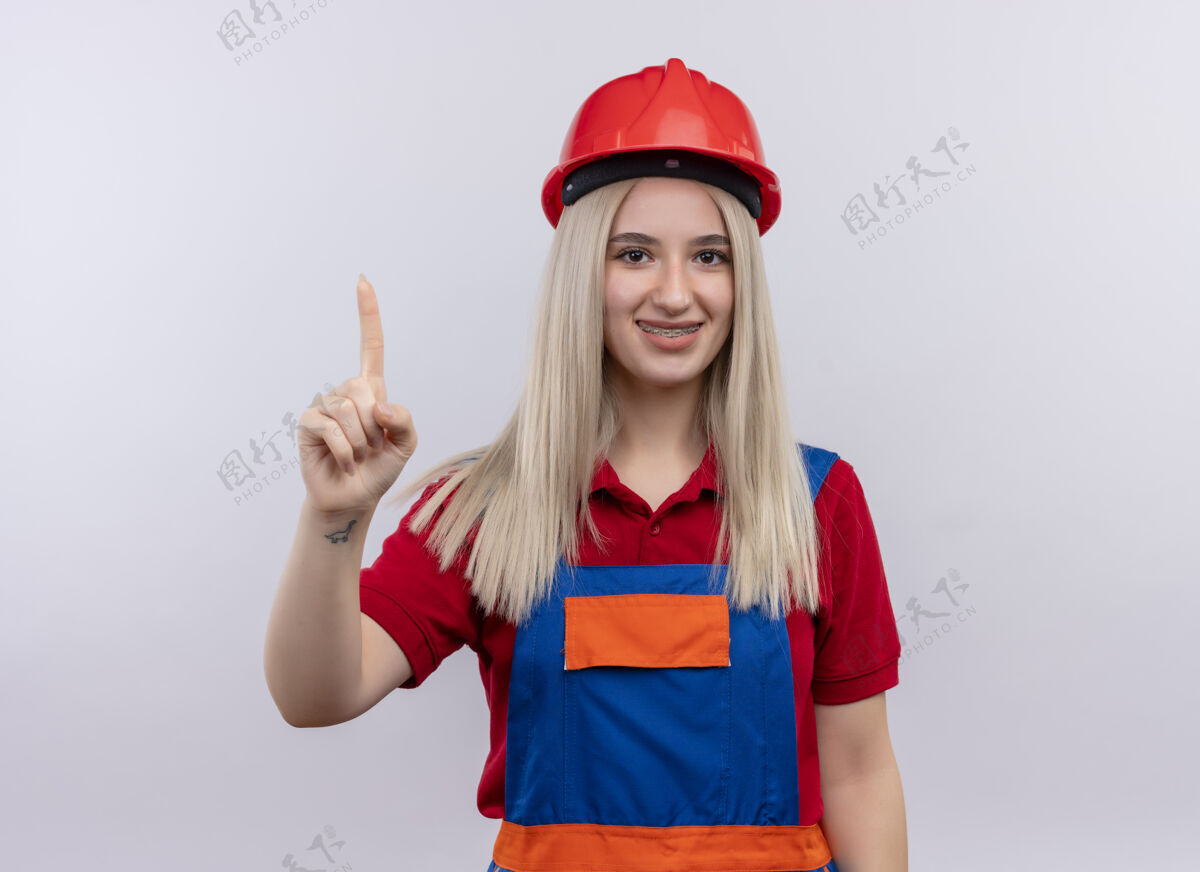 举起来微笑着的年轻金发工程师建筑工女孩穿着制服 戴着牙套 在孤立的空白处竖起手指年轻人牙套手指