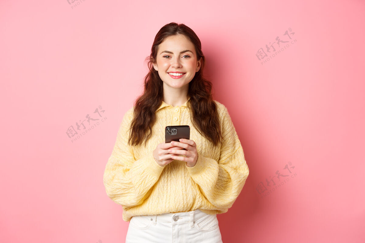 快乐手持手机微笑的美丽女孩的肖像 网上购物 使用社交媒体应用程序 穿着休闲服站在粉红色的墙上休闲女性手势