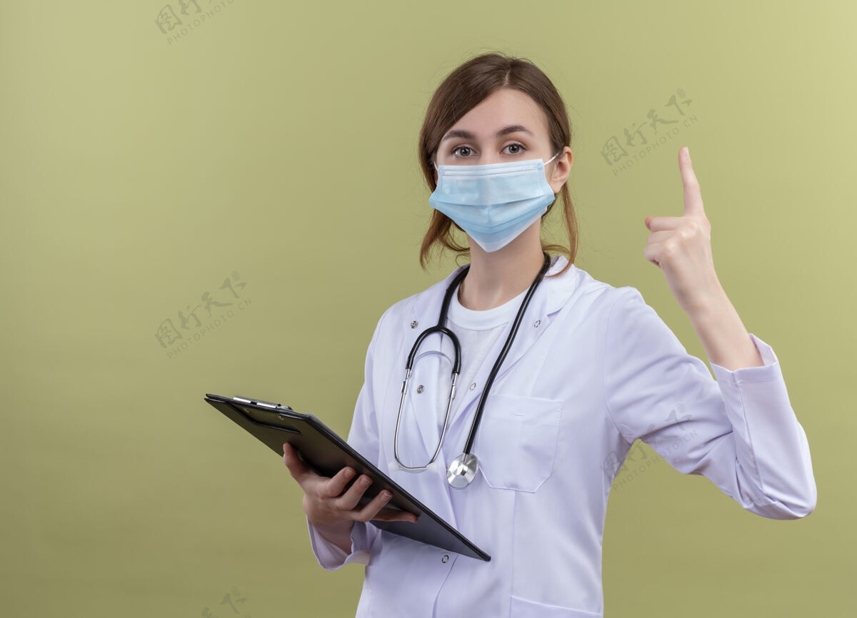 医生面容严肃的年轻女医生 穿着医用长袍 戴着口罩 手持听诊器 指着隔离的绿地绿色向上年轻