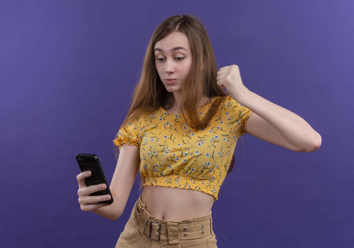 女孩神情严肃的年轻女孩拿着手机 在孤立的紫色空间举起拳头严肃年轻拳头