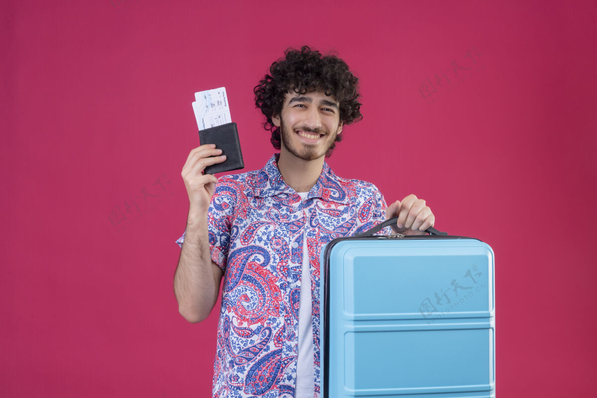 男人快乐的年轻帅气的卷发旅行者手里拿着钱包 机票和手提箱 放在粉红色的空间里 还有复印空间手提箱机票钱包