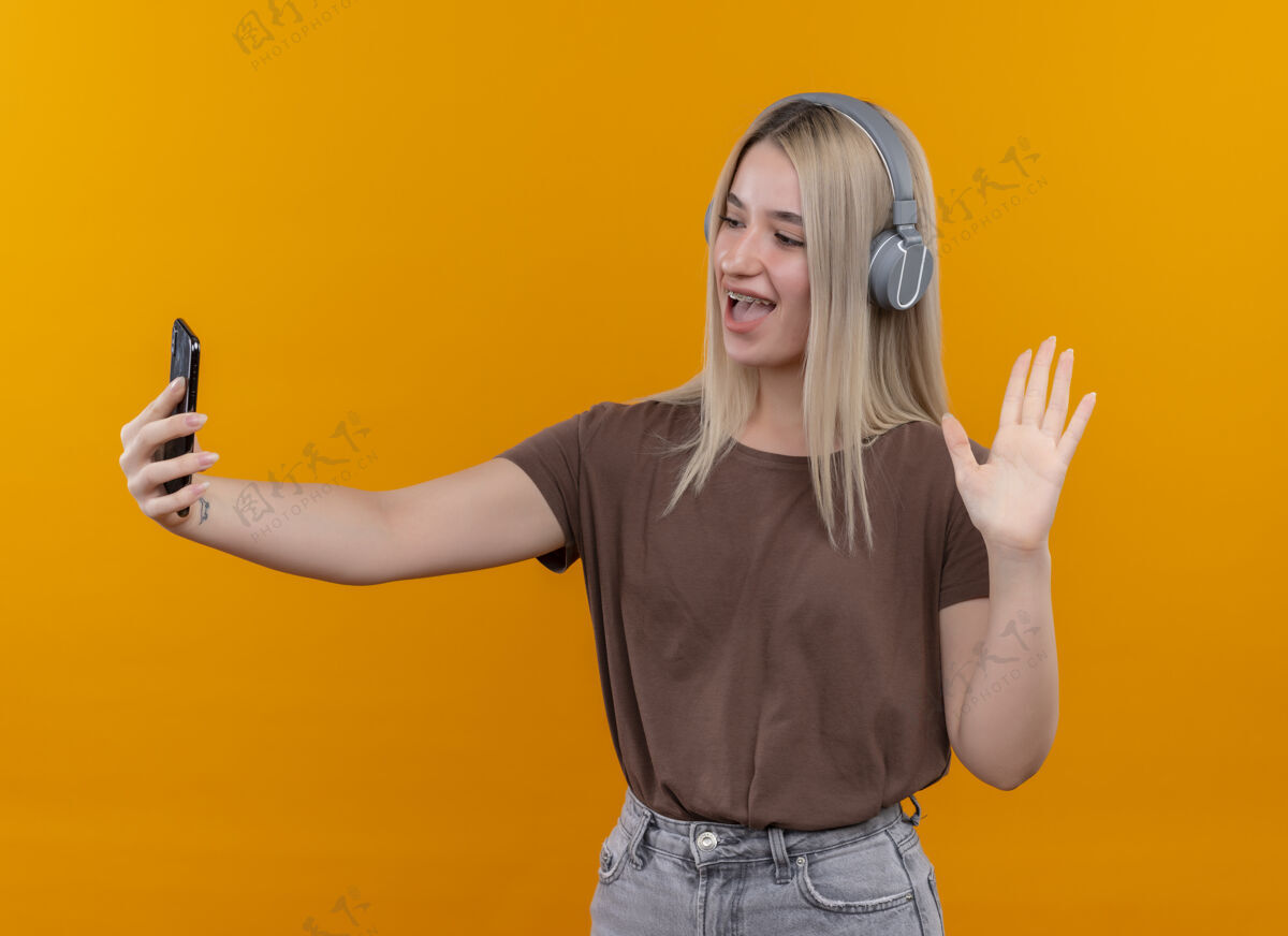 金发快乐的年轻金发女孩戴着耳机戴着牙套拿着手机看着它在孤立的橙色空间挥手手机年轻喜悦