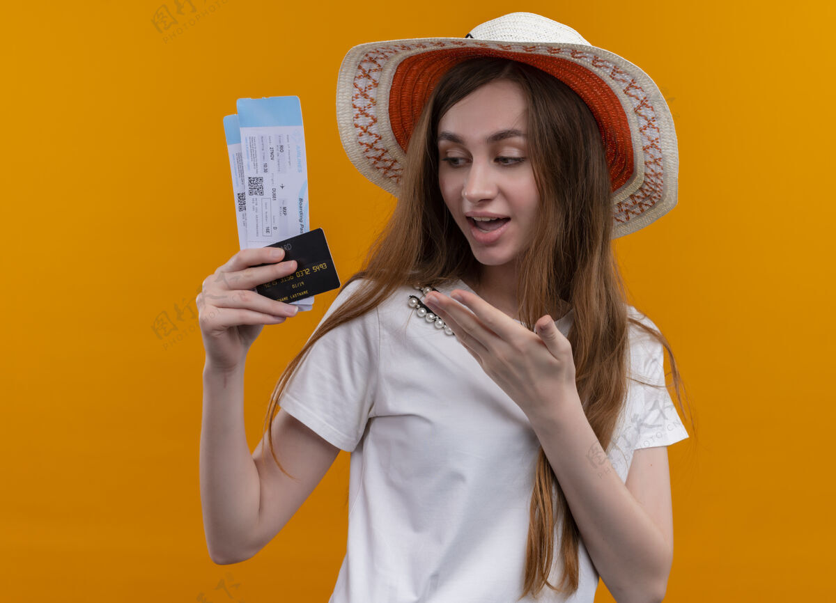 机票令人印象深刻的年轻旅客女孩戴着帽子拿着机票 信用卡指着他们用手在孤立的橙色空间飞机女孩年轻