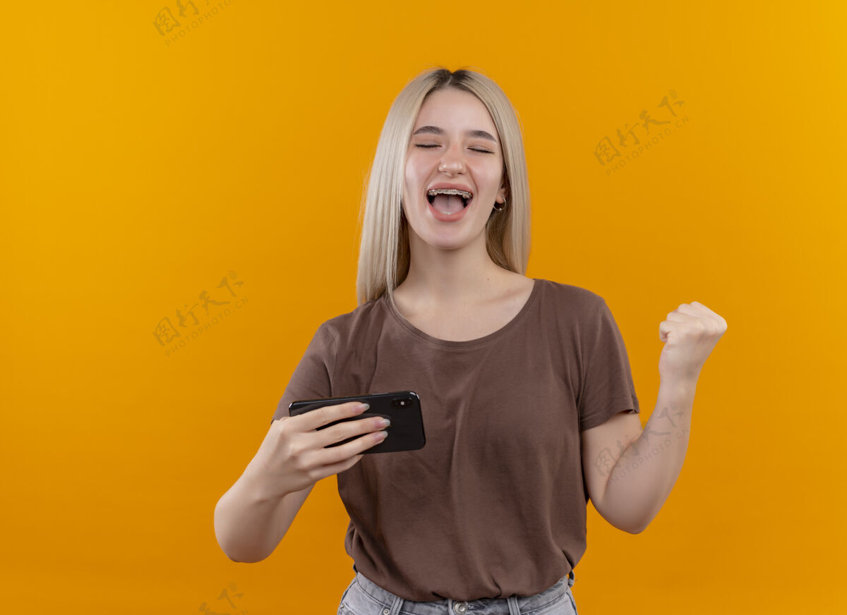 金发戴着牙套的快乐的金发女孩举着手机 举起拳头 闭上眼睛 看着孤立的橙色空间和复印空间眼睛橙色复制