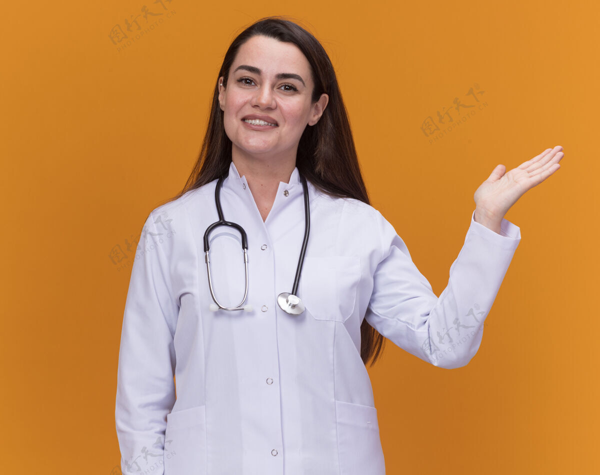 年轻微笑着的年轻女医生穿着带听诊器的医用长袍 手张开放在橘子上手听诊器医生