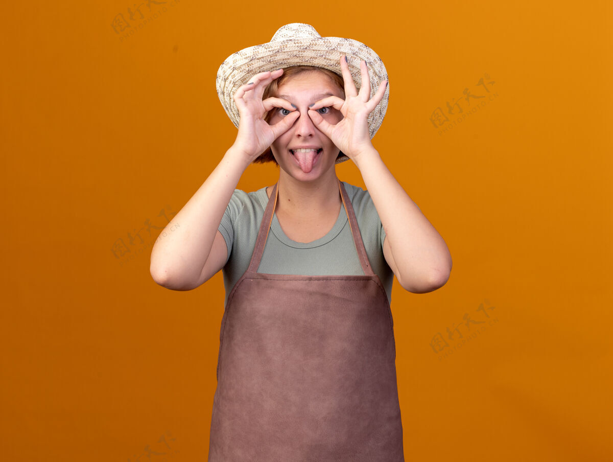 通过有趣的年轻斯拉夫女园丁戴着园艺帽 结结巴巴地伸出舌头 用手指盯着相机看橙色年轻园艺卡住