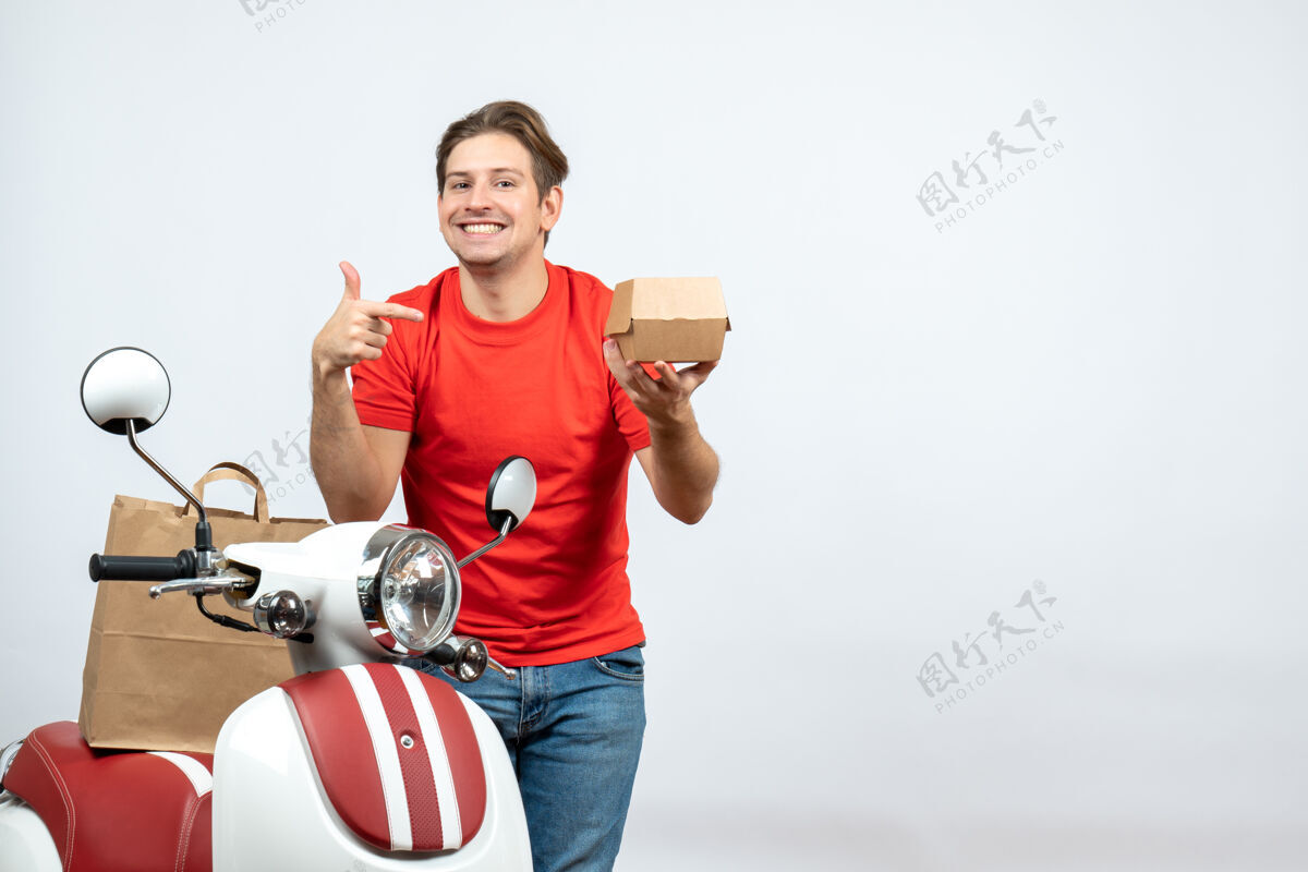 男性俯视图中微笑的送货员穿着红色制服站在滑板车旁 指着白墙上的订单快乐送货员男人