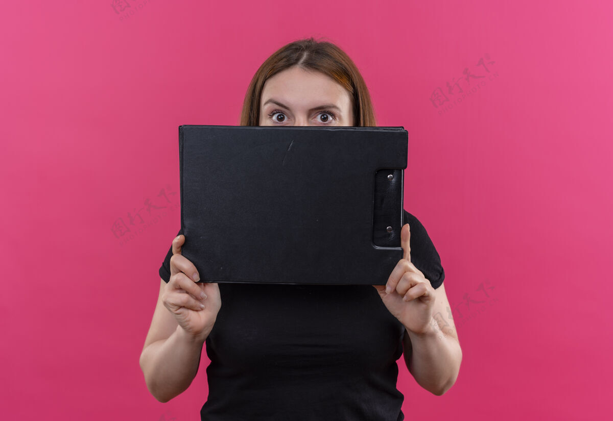 粉红印象深刻的年轻随意的女人拿着剪贴板 躲在它后面的孤立的粉红色空间剪贴板女人隐藏